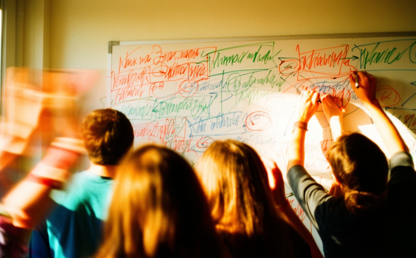 A whiteboard dense with coloured handwriting and arrows, two people writing at once.
